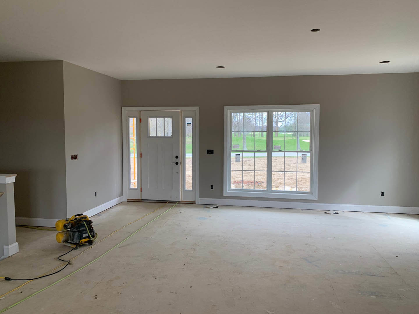 White-walled room with yellow and black air compressor, yellow wires along baseboard, white door with glass panes, window overlooking golf course, white pillar beside brown door