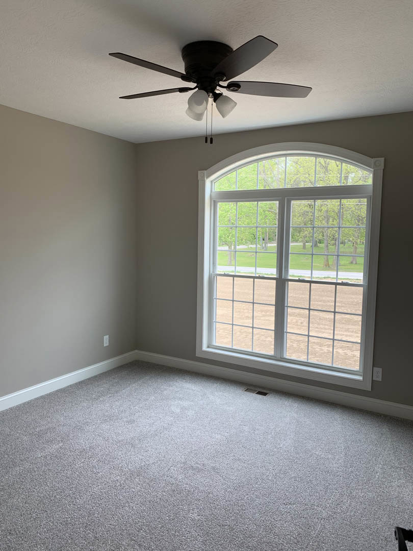 Ceiling fan with integrated light fixture mounted on white ceiling above carpeted floor, large window offering view of trees and bare ground.