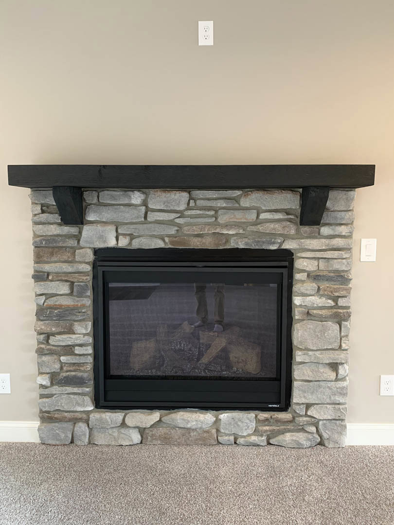 Stone fireplace with black metal frame and hearth, wood-burning stove, carpeted floor, and black shelf above stone wall in living room.