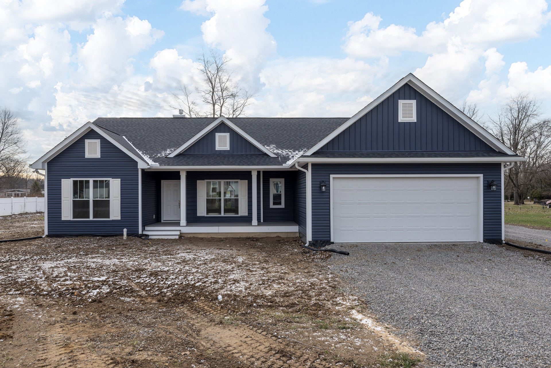 Blue siding house with white garage door, gravel driveway, white-framed window, black roof, white vent, and tree under cloudy sky