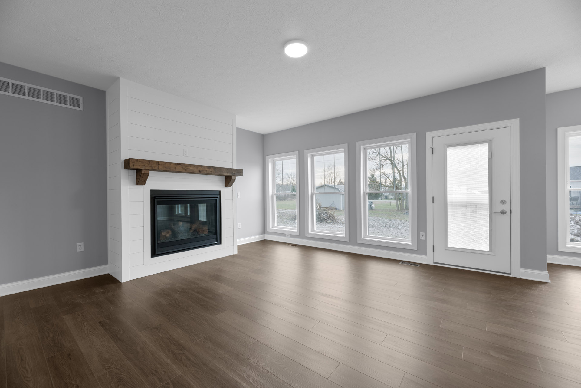 Living room with wood flooring, plaster walls, ceiling light, fireplace with glass door, multiple windows with blinds, and a white door featuring a window.