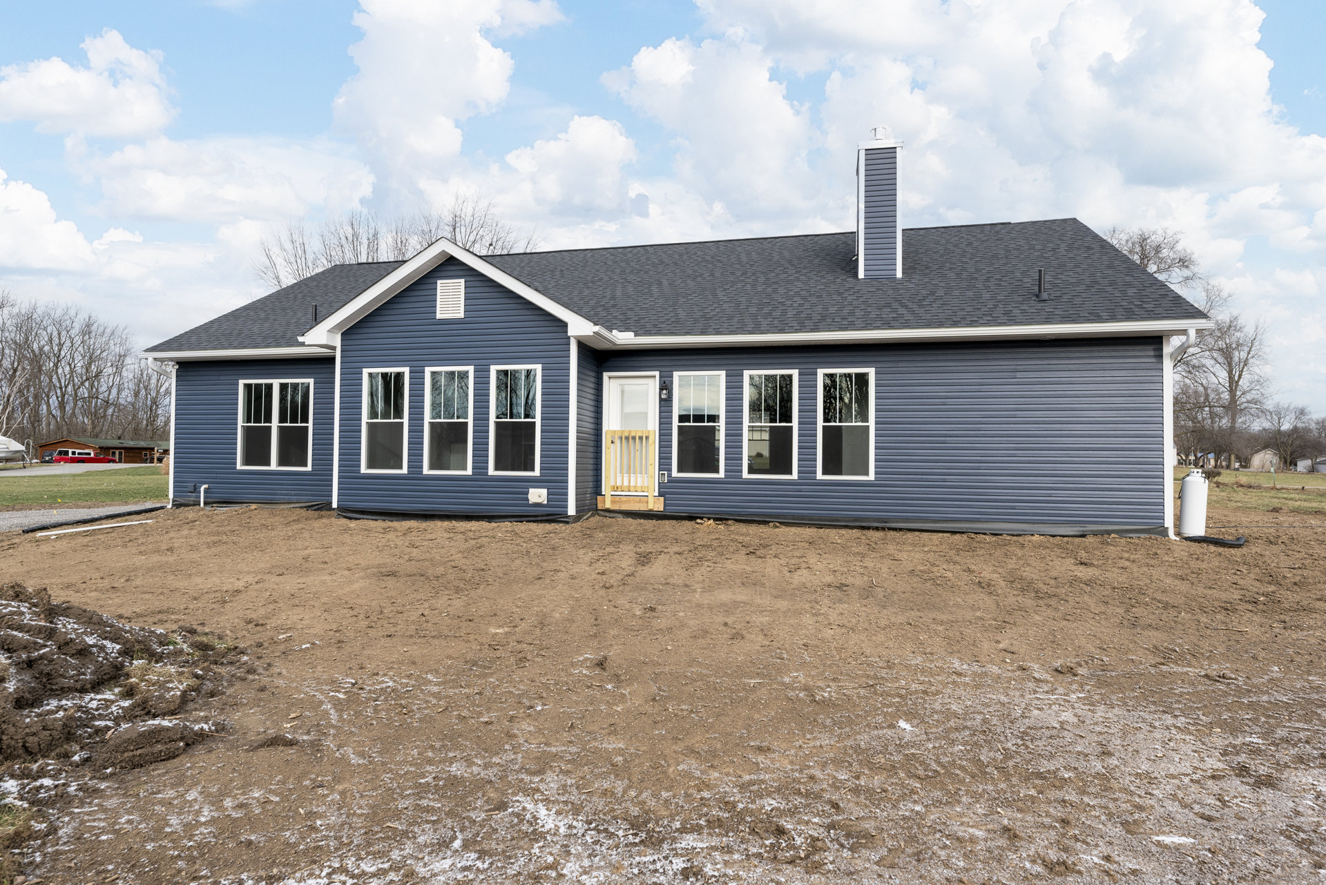 Two-story house with white-framed windows, wooden front door with glass panel, situated on a dirt hill with sparse grass, tree reflected in window, cloudy sky overhead