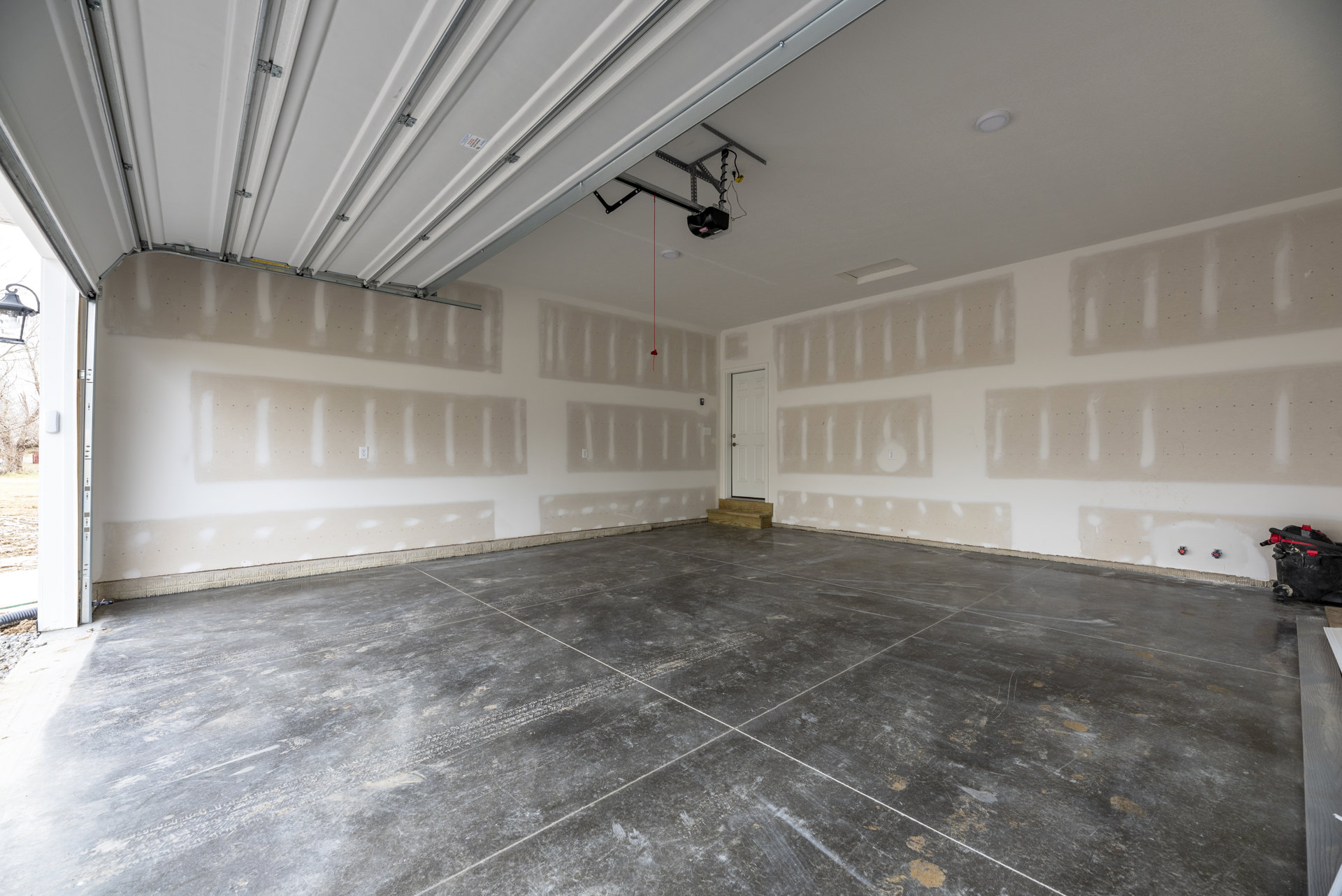 Concrete floor with white tile lines, white plaster walls, ceiling, garage door, and a white door featuring a glass handle; black and red vacuum on floor.