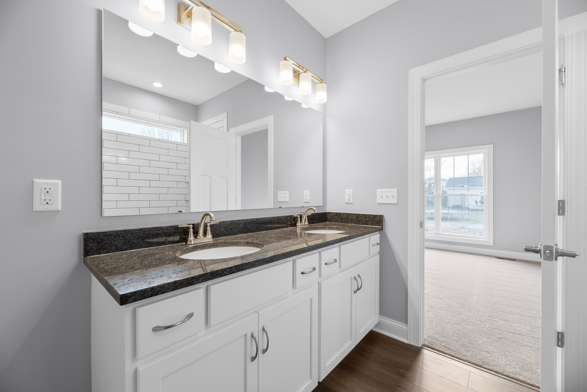 Bathroom with white shaker cabinets, matte black countertops, chrome faucet, tile flooring, wall-mounted light fixture, white outlet, light switch, window overlooking backyard