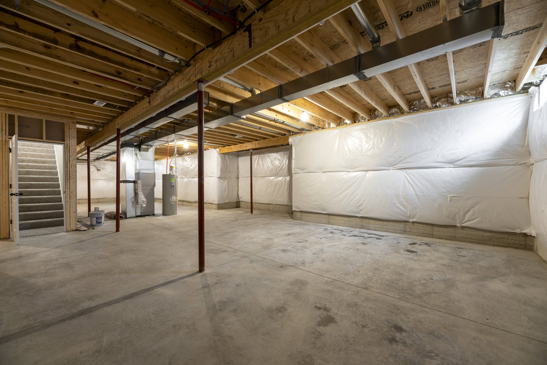 Spacious unfinished room with a large white wall, concrete floor, red support pole, staircase leading to a door, wall covered in plastic, and paint bucket on the floor