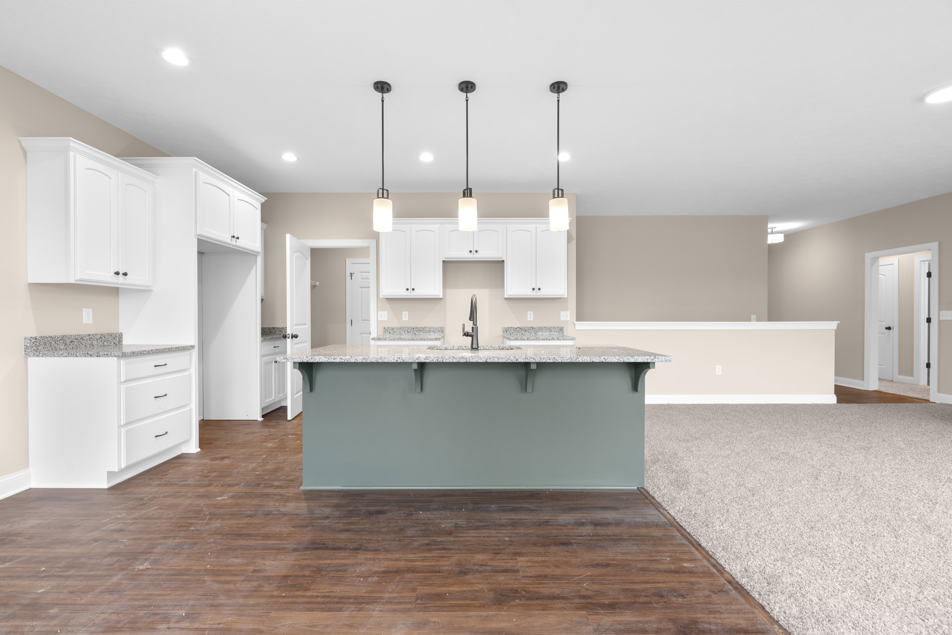 Spacious kitchen featuring a large central island with built-in sink and faucet, wood flooring painted white, white cabinetry, black hardware, and a carpeted area adjacent to a