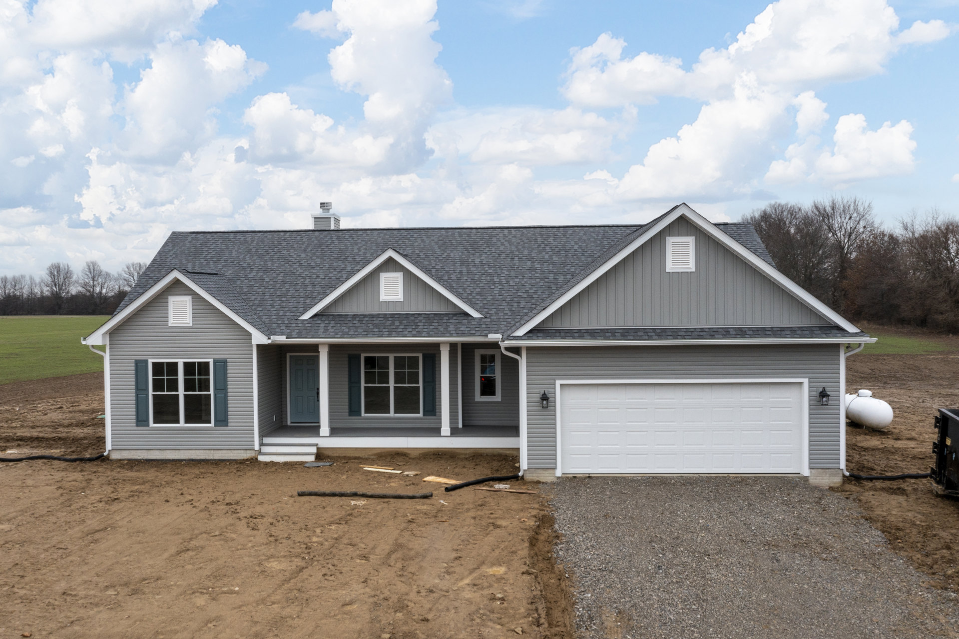 Grey siding custom home with white-framed windows, white garage door, grey roof, brick chimney, paved driveway, and cloudy sky backdrop