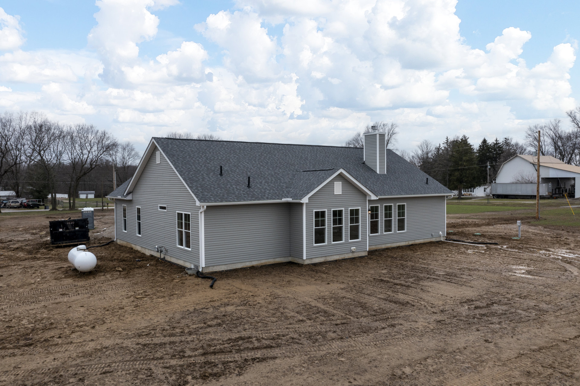 Grey house with white roof under construction on dirt lot, black dumpster with numbers in foreground, white cylinder on ground, scattered trees and clouds in sky