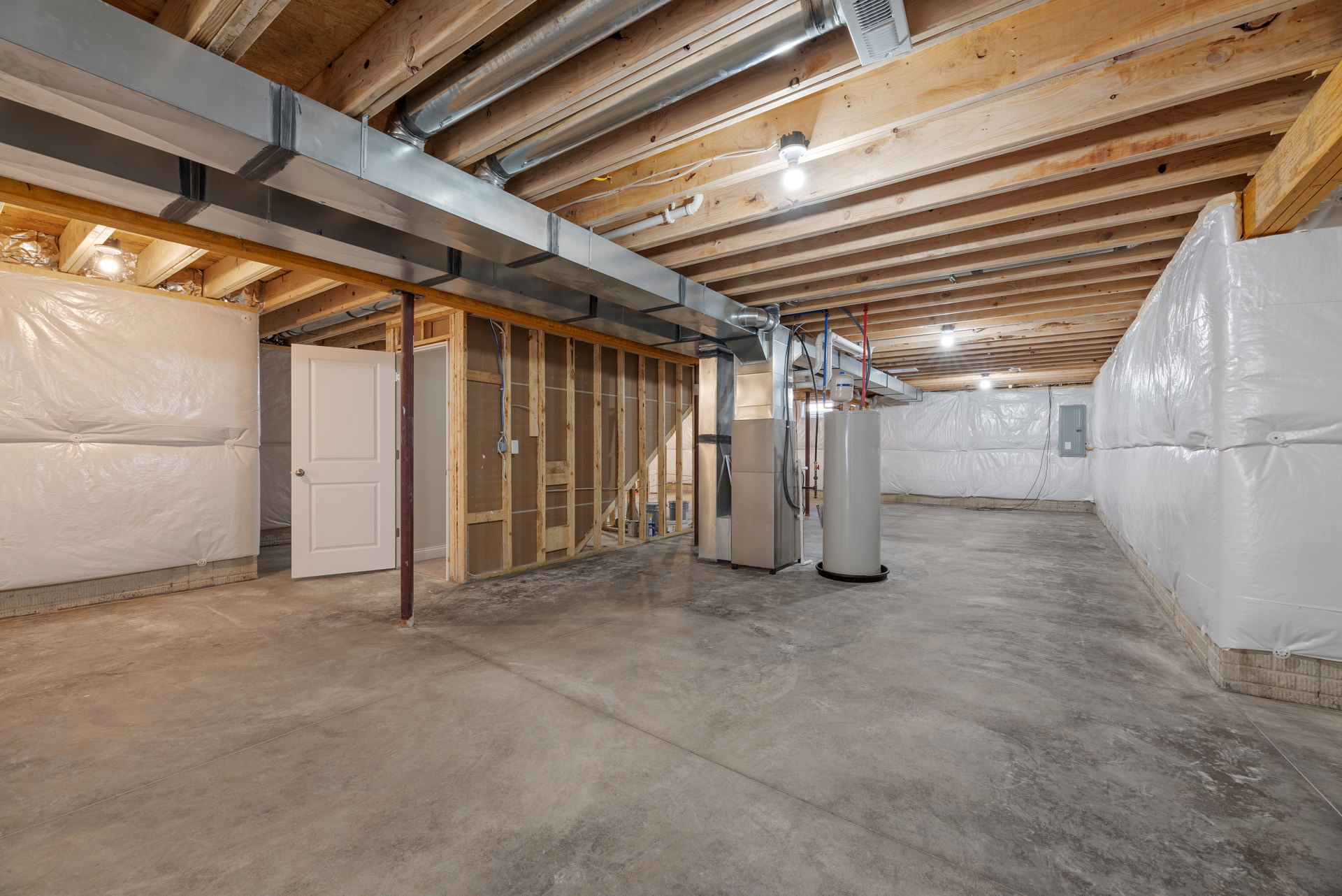 Basement room with exposed wood beams and ceiling, concrete floor, white cylindrical heater with metal pipe, white door with silver handle, structural column, wooden framing, white