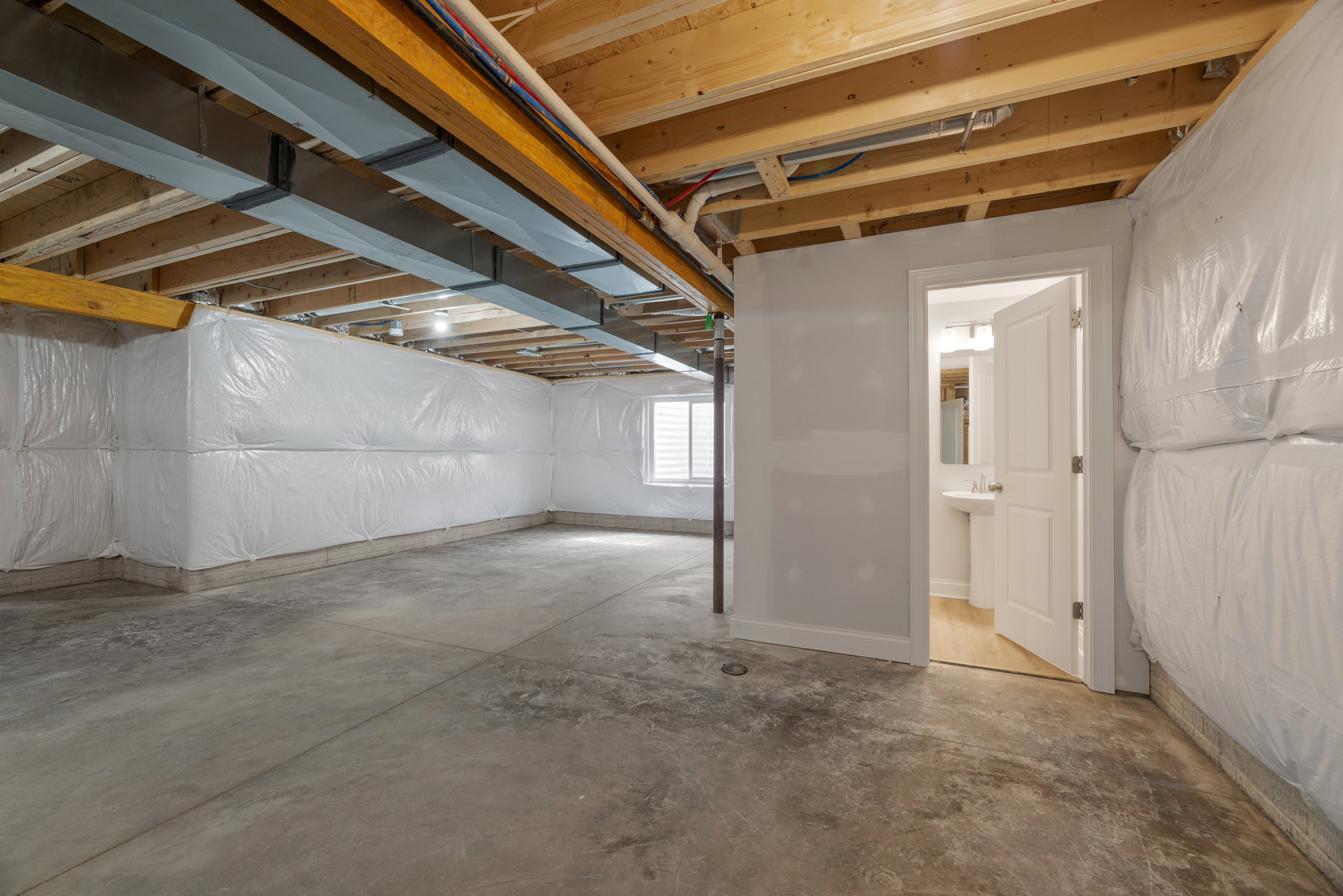 Bathroom with white plaster walls, concrete floor, open door, exposed metal beams and pipes on wood ceiling, wall-mounted sink, and rectangular mirror.