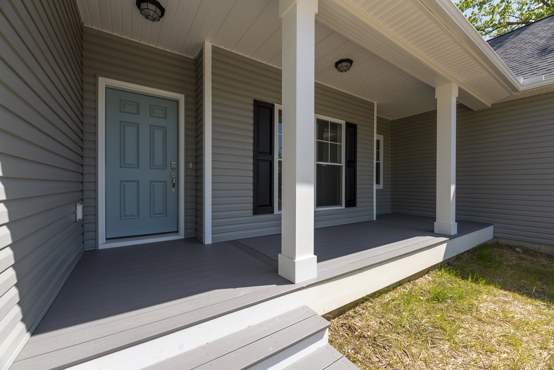 Front porch with white siding, grey door featuring a lock, black shutter, white-framed window, and decorative lamp above entry; manicured grass borders the house.