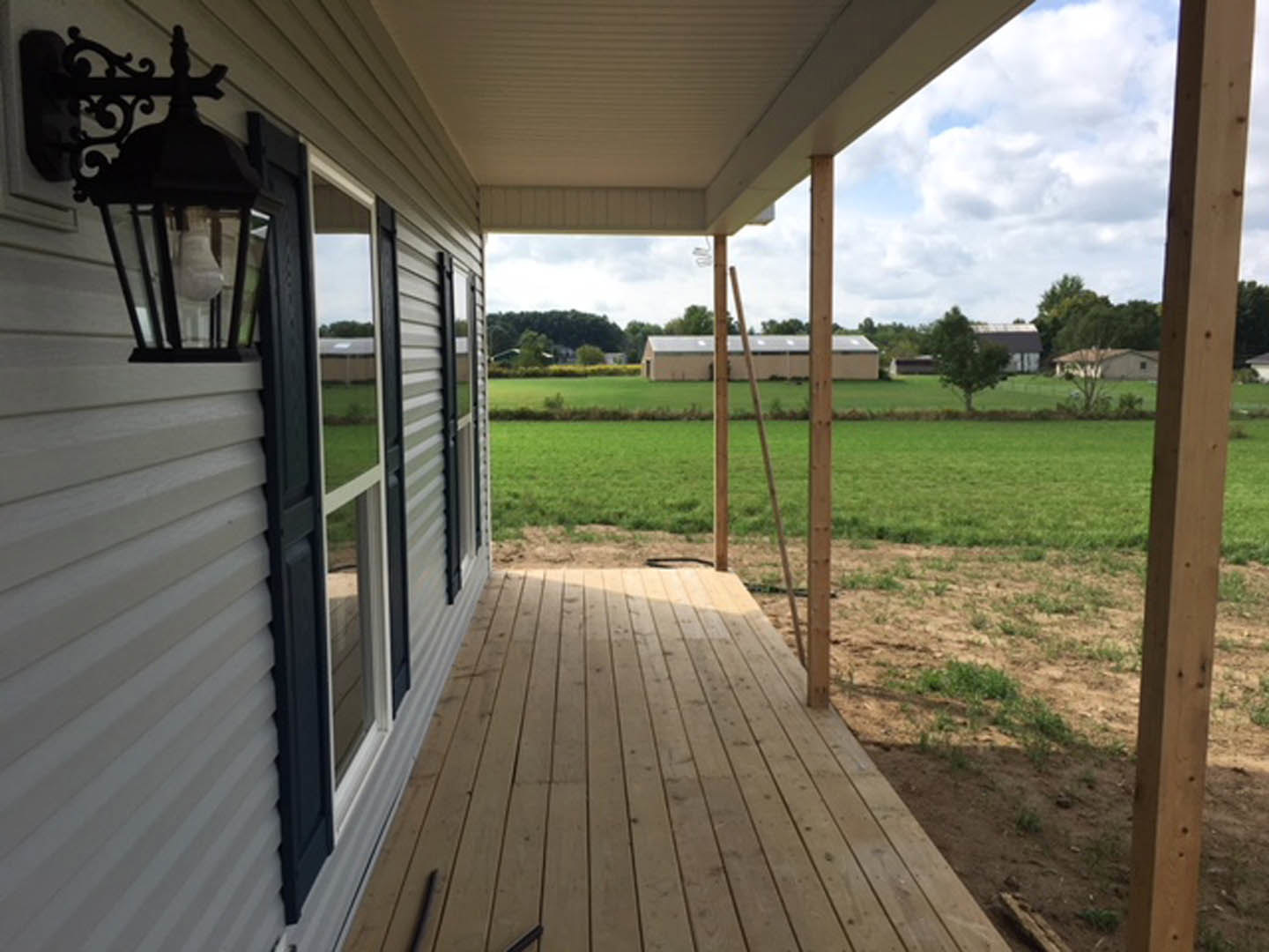 Modern home with large windows, wooden deck and fence, surrounded by a field of green grass and scattered trees under a blue sky
