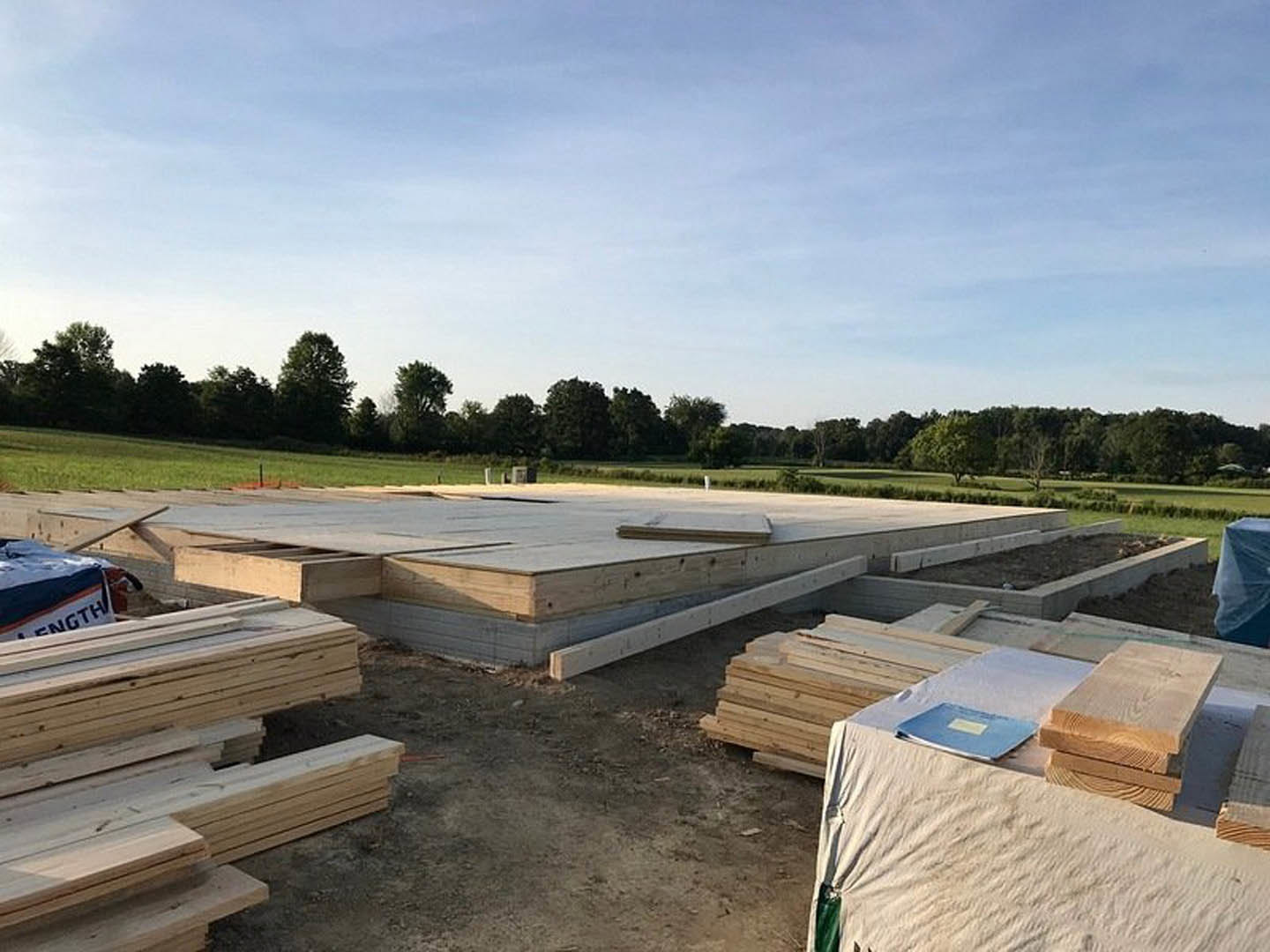 Stack of lumber planks on dirt ground at residential construction site, blue sky and scattered clouds overhead, grassy area and partially built structure in background.