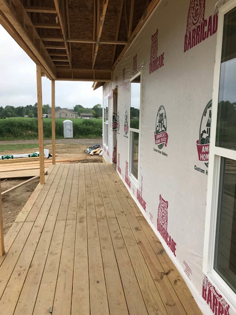 Covered porch with wooden deck, white exterior wall, exposed wooden beams, large window, and red logo sign.