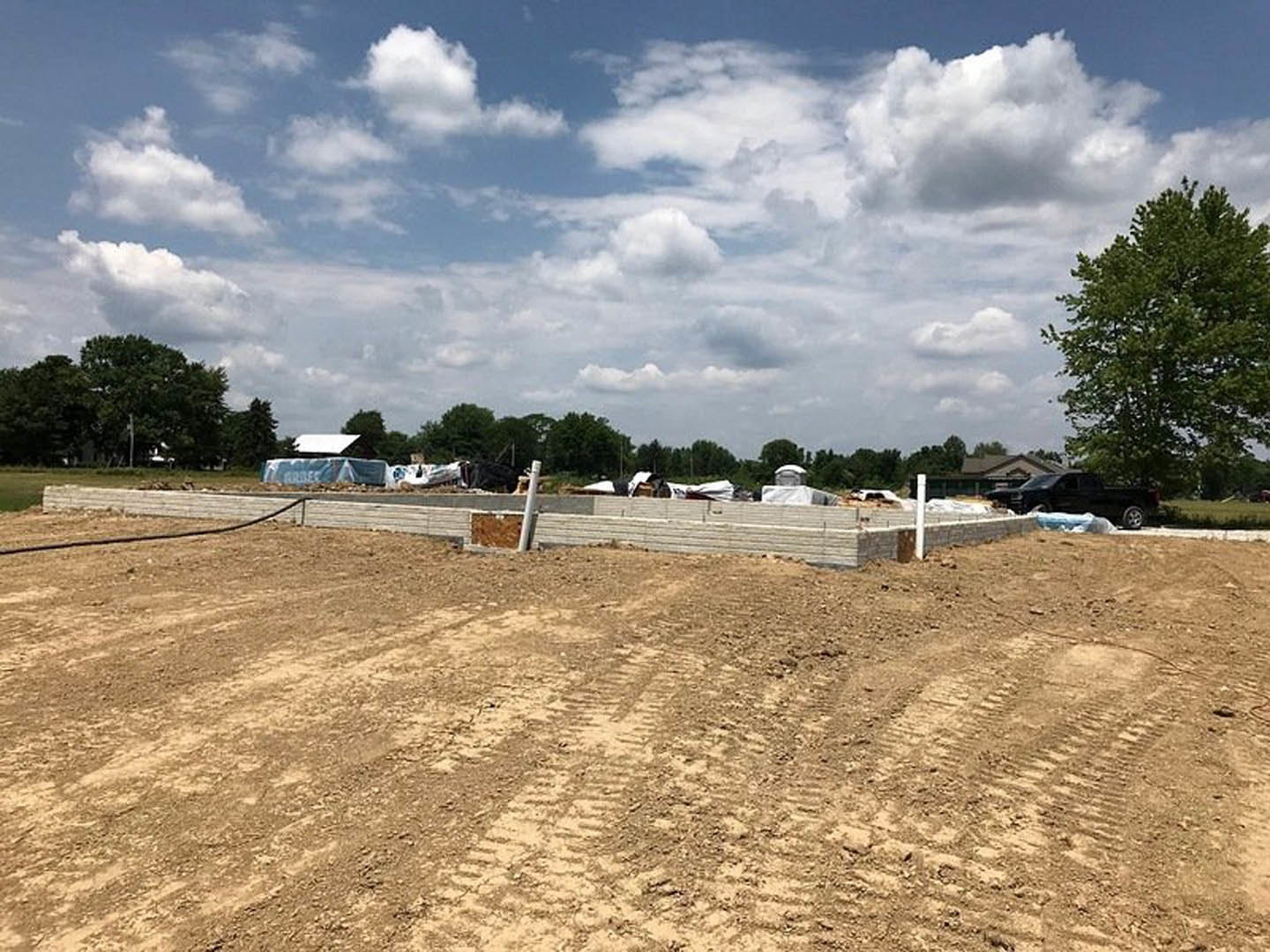 Dirt construction site with tire tracks, leafy trees, cloudy sky, black truck parked near partially built house, close-up of concrete wall