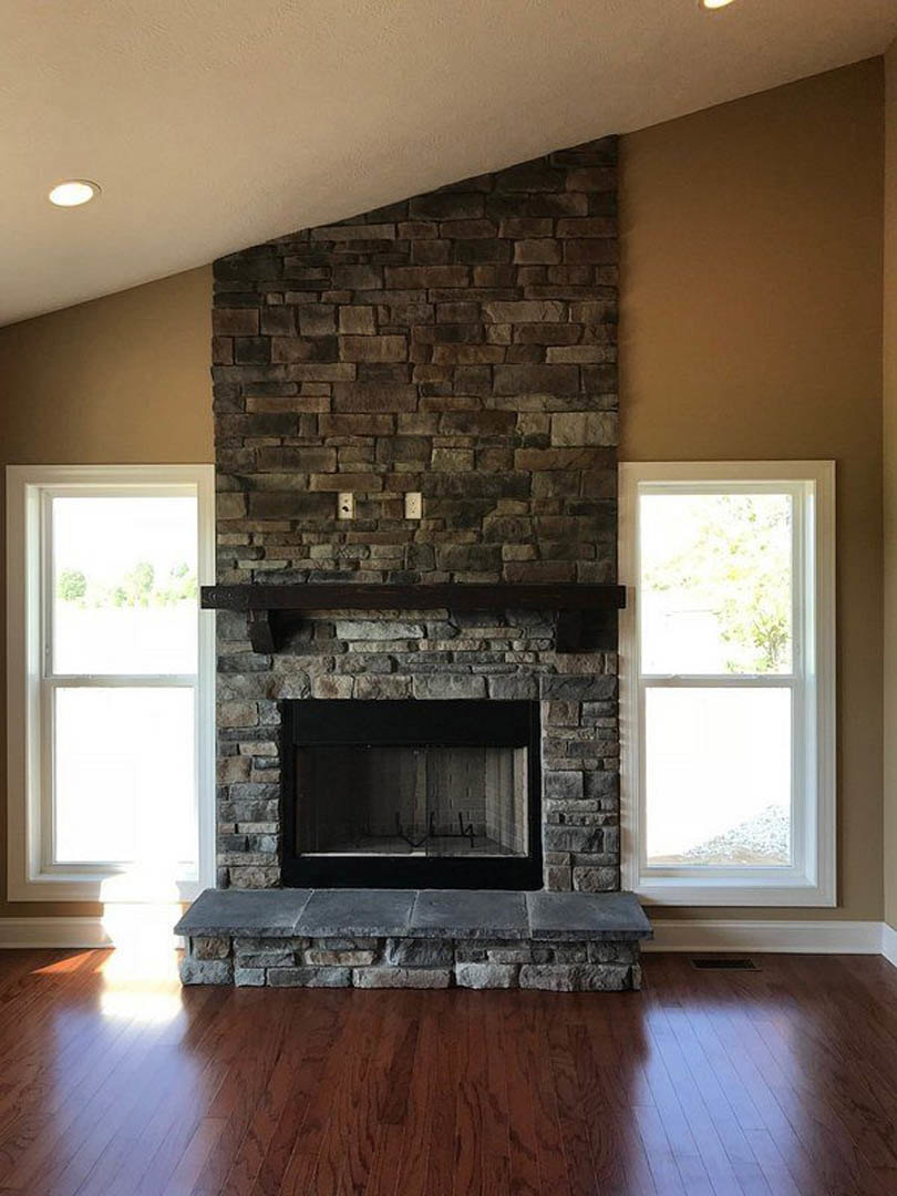 Stone fireplace with glass window and wood mantel, white-framed window, wood flooring, and fire screen in a cozy living room.