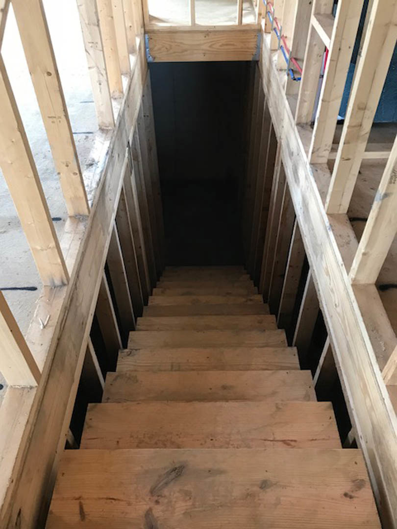 Wooden staircase with matching handrail ascending in a narrow, dimly lit interior, natural light streaming through a nearby window, highlighting wood plank flooring and exposed