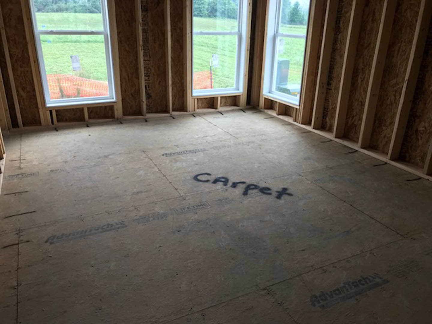 Wood-framed room under construction with large windows, exposed beams, and subfloor marked with black text, overlooking grassy field.