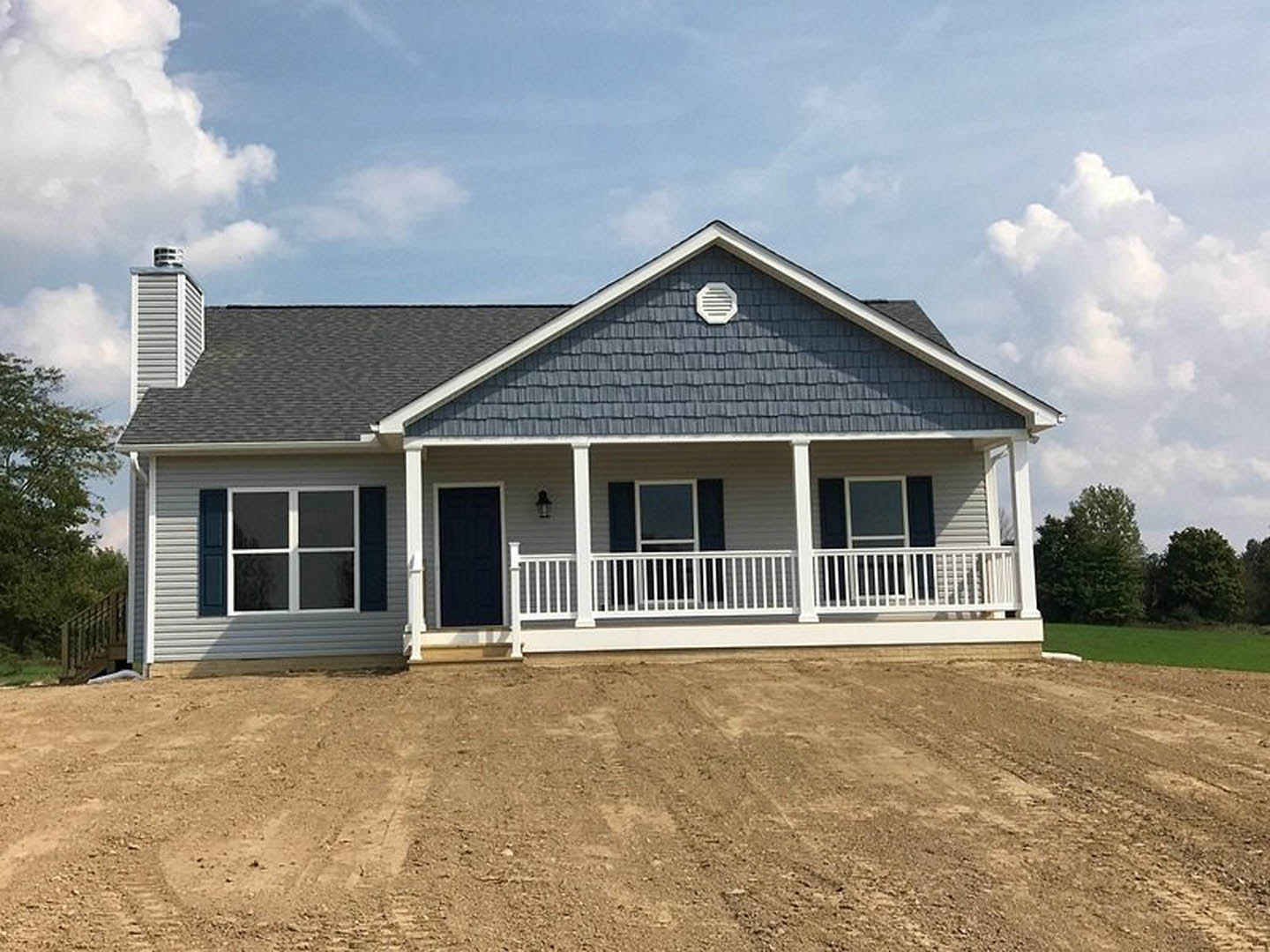 Two-story house with black door and white trim, covered porch, white vent on exterior wall, white-framed windows, dirt hill and field bordered by white railing, cloudy sky