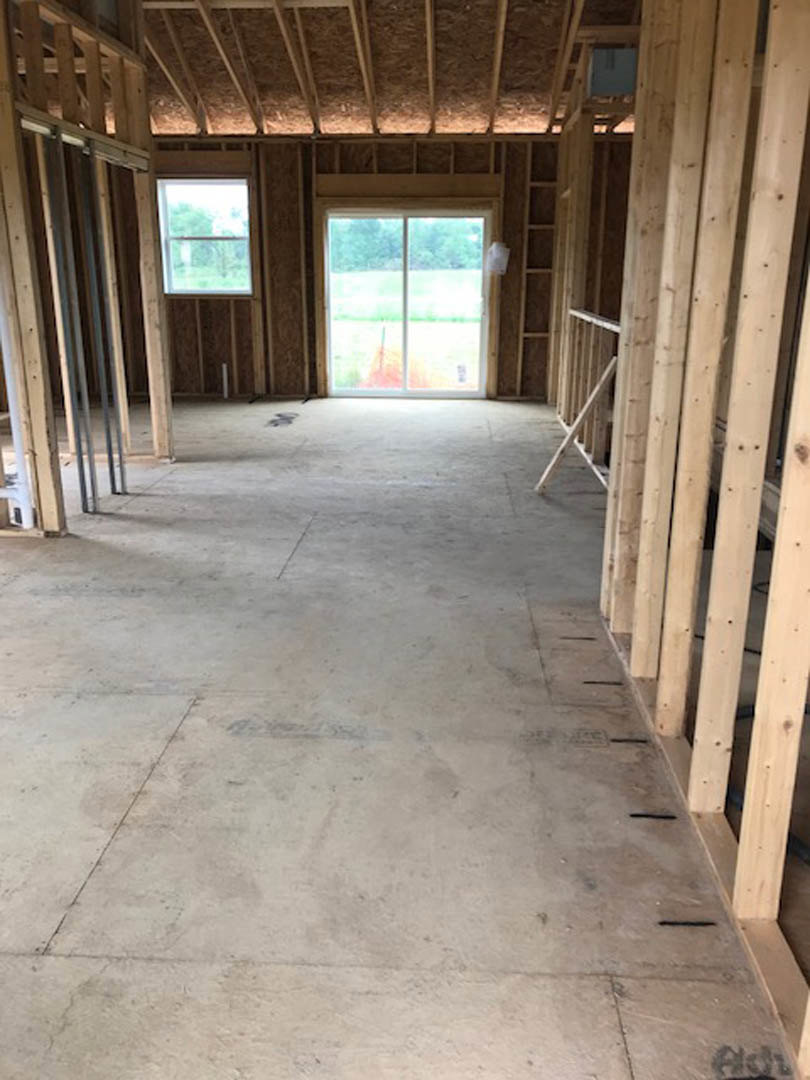 Bright room with wide-plank wood flooring, white railing, large window overlooking a green field and trees, upholstered chair in foreground