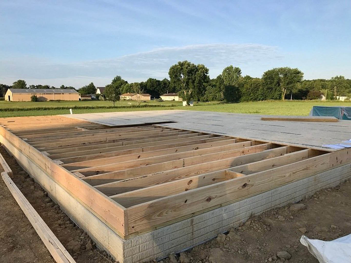 Wooden house frame under construction with exposed beams, foundation, and grassy outdoor setting; tree and cloudy sky in background.