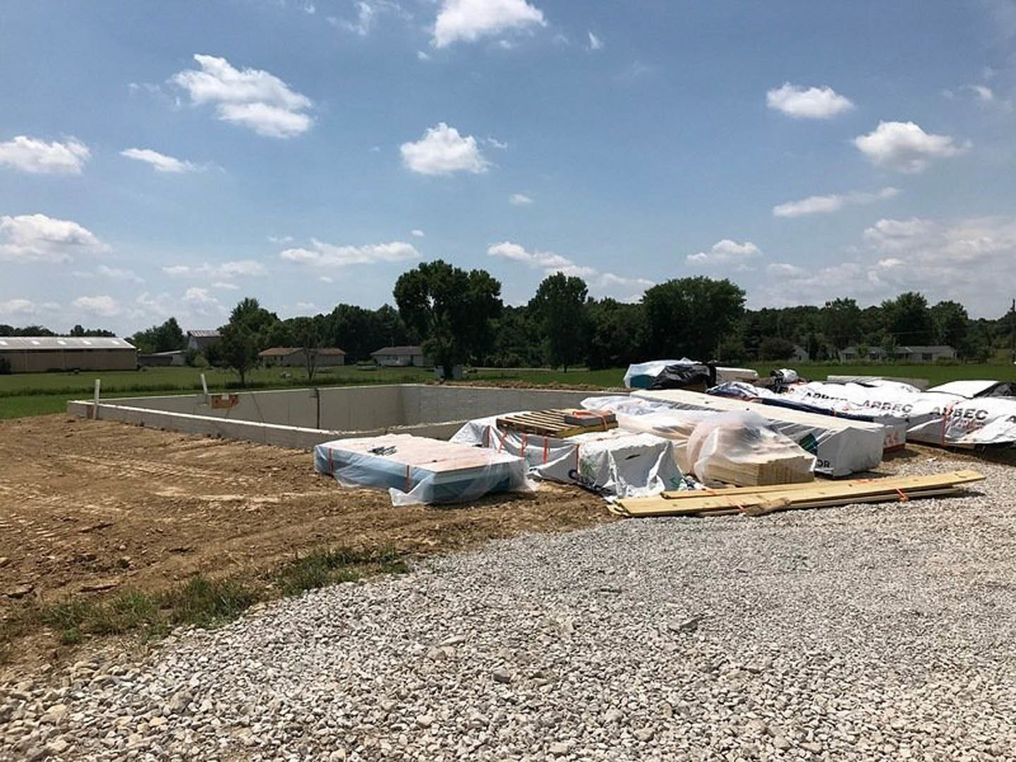 Concrete foundation surrounded by construction materials, gravel, wooden planks, and a mattress on the ground; partially built structure with roof in background, blue sky and trees