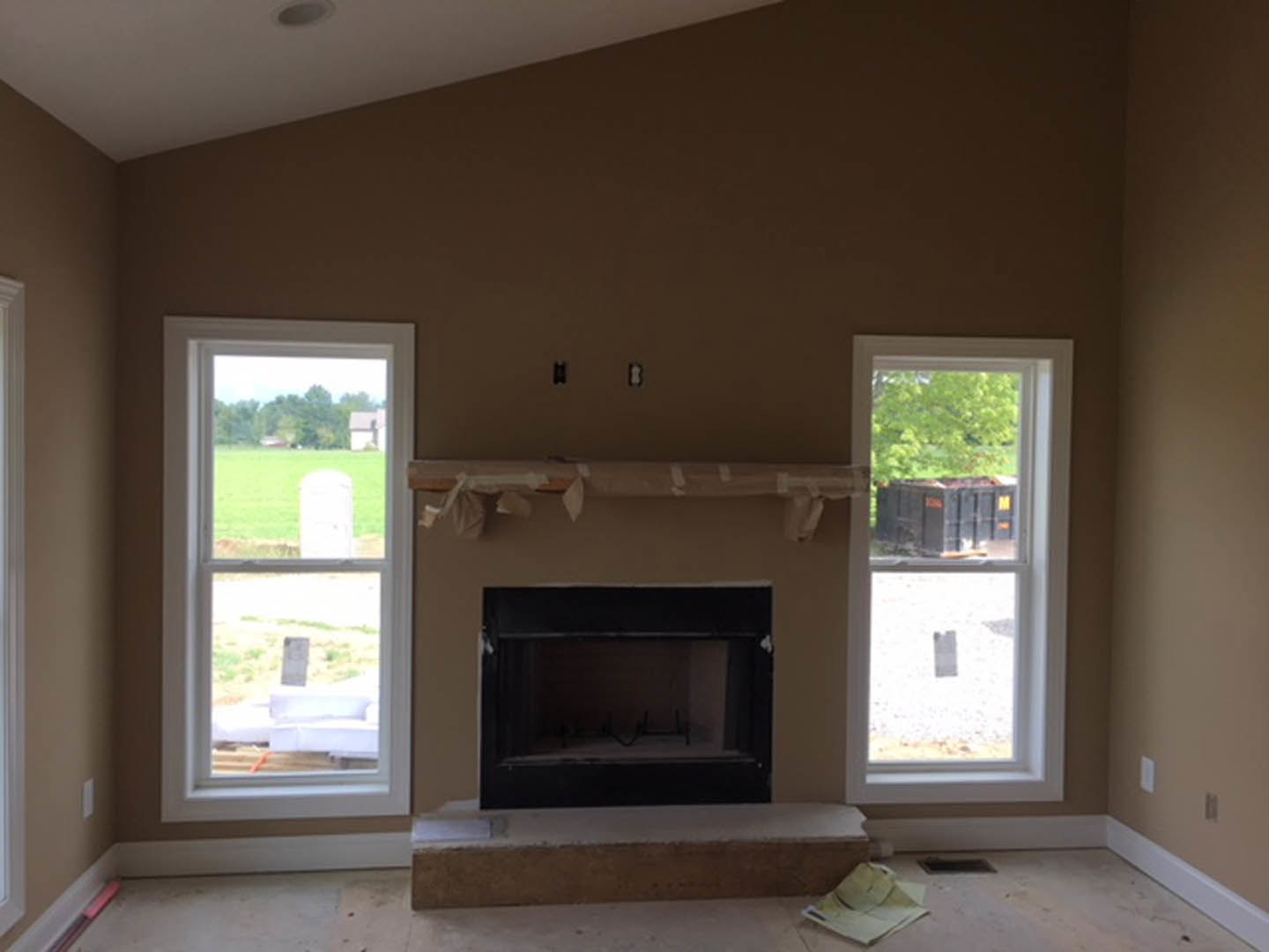 Stone fireplace with white mantel in a cozy living room, hardwood floors, large window overlooking grassy field, wooden table with hat, neutral walls