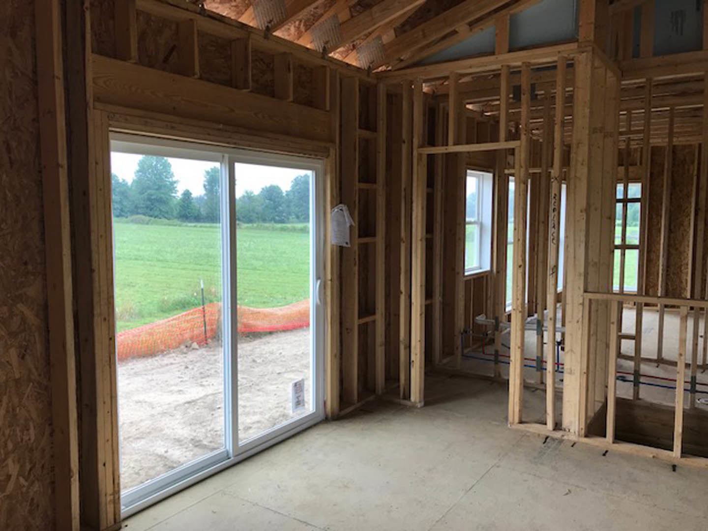 Open room with exposed wooden beams, concrete floor, sliding glass door overlooking grassy field, large window, and wood surfaces.