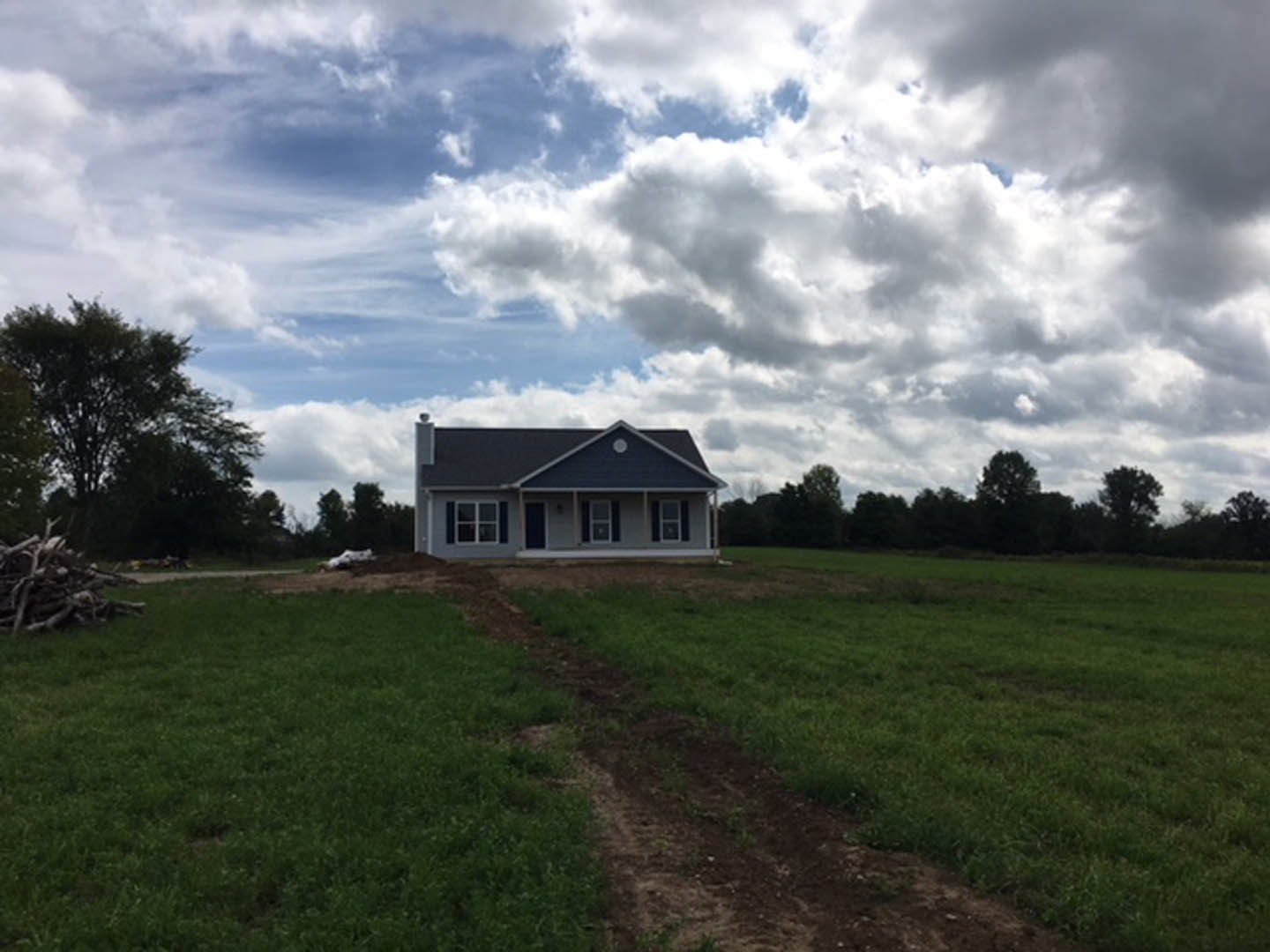 Modern farmhouse with gabled roof, wood siding, and large windows, surrounded by grassy field, mature trees, and a dirt path leading to the entrance under a partly cloudy sky