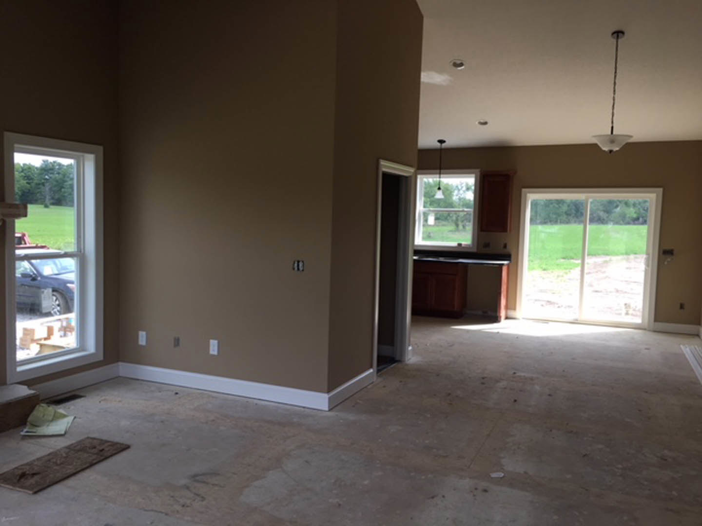 Open white door leading to a bright room with light wood flooring, large window overlooking grassy field and parked car, smooth plaster walls, and minimal interior fixtures.