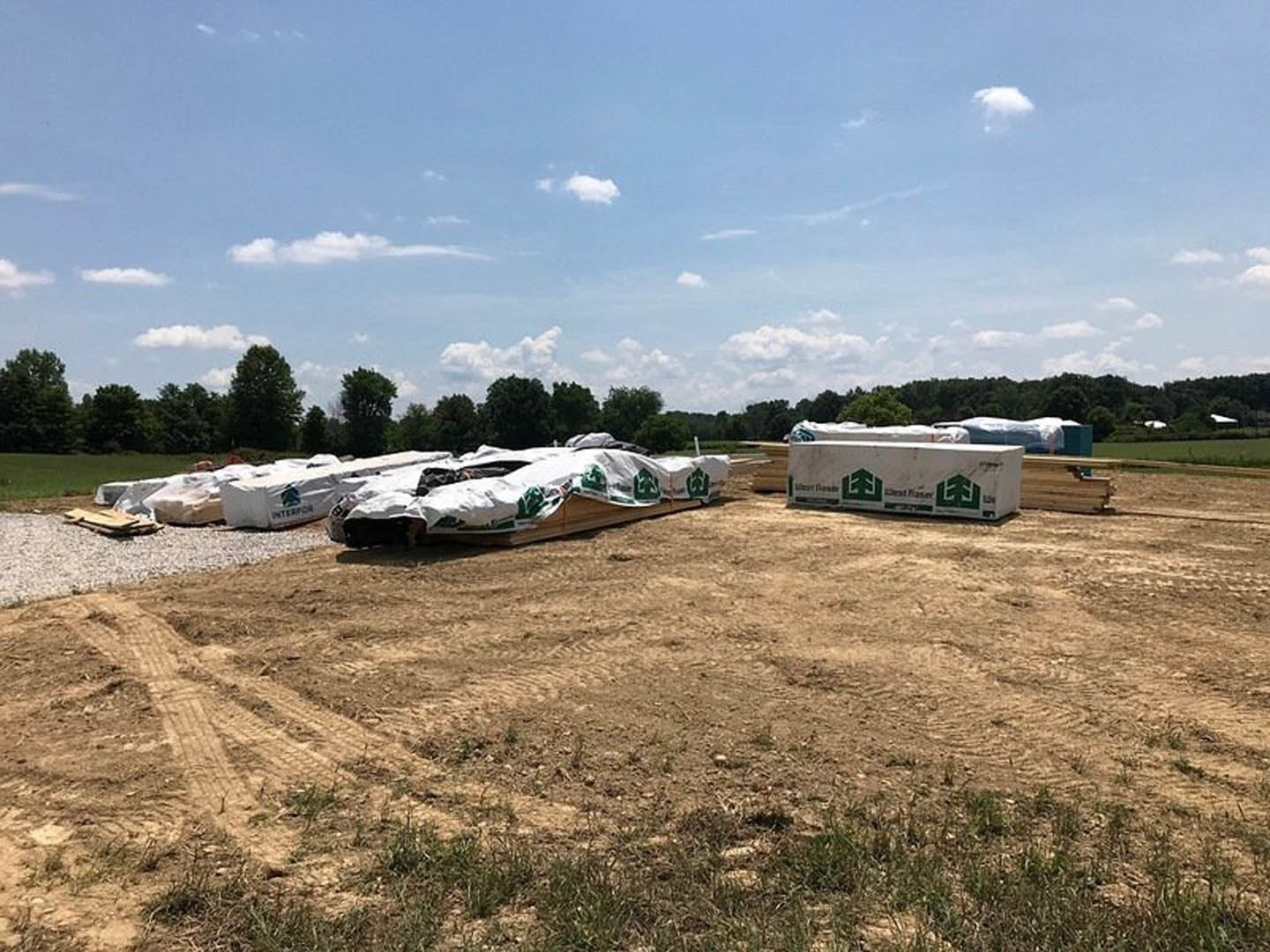 Wide grassy field with scattered white tarps, mature trees in the background, dirt area with stacked wood, blue sky with clouds overhead, white tarp covering a wooden box, green