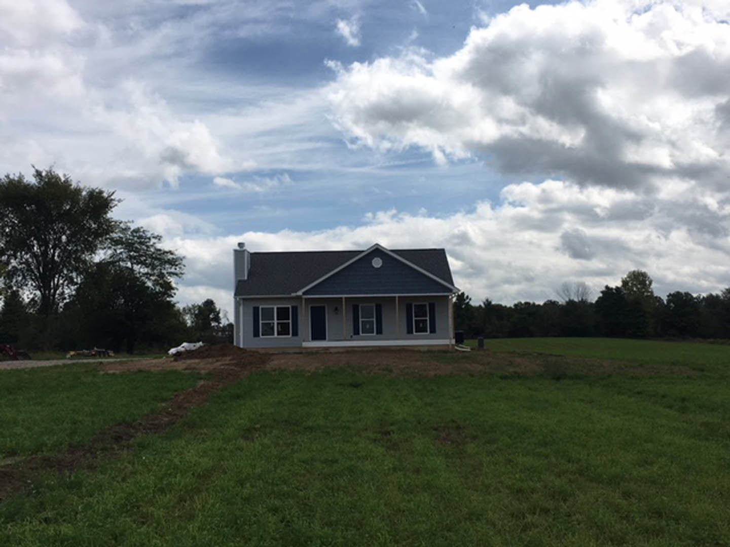 White custom home with gabled roof and large windows, surrounded by green grass field, mature trees, and cloudy sky in rural setting