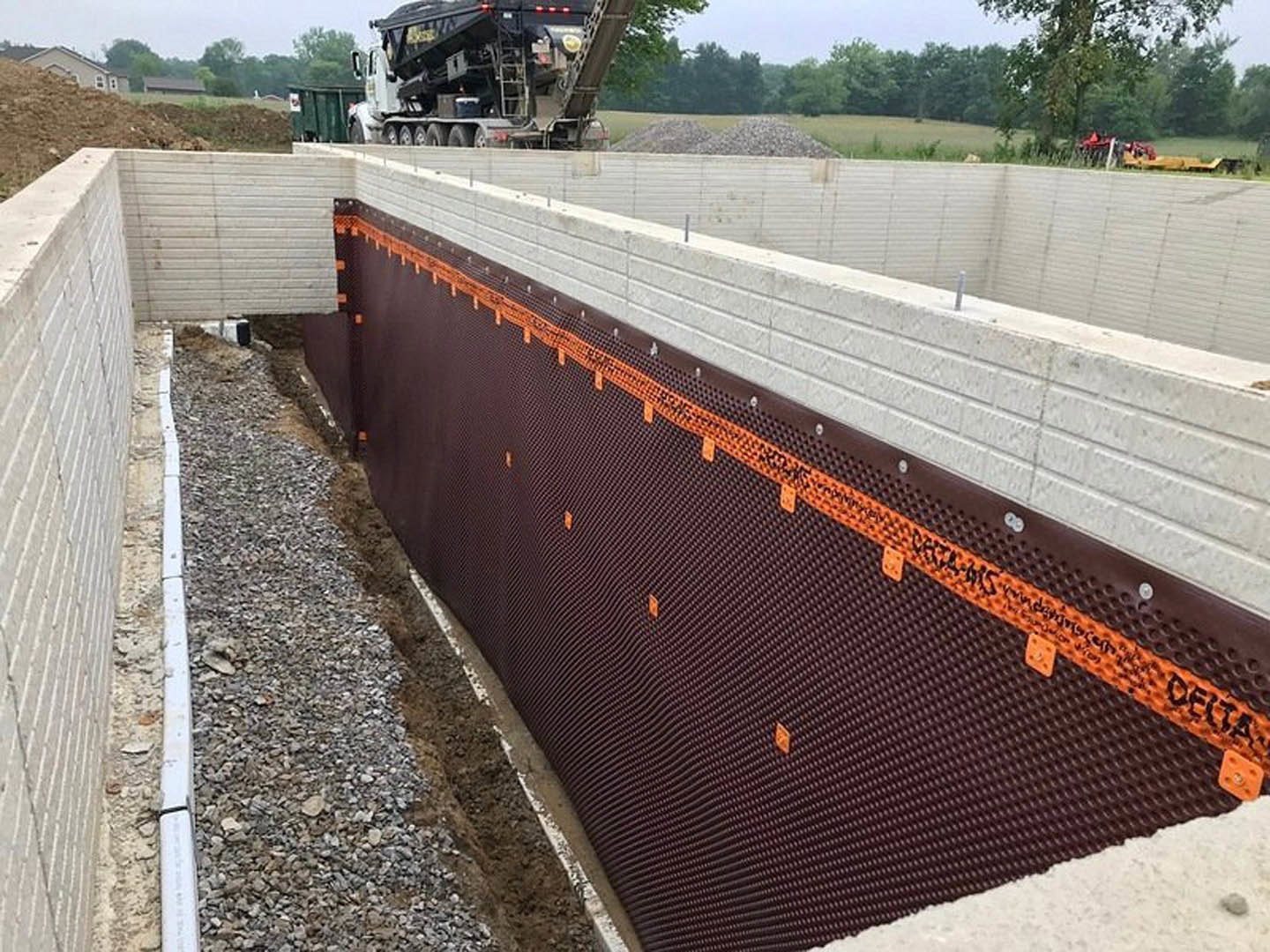 Gravel construction site with partially built wall marked by orange tape, large black truck with ladder parked near white line, fenced perimeter, and field of trees in background