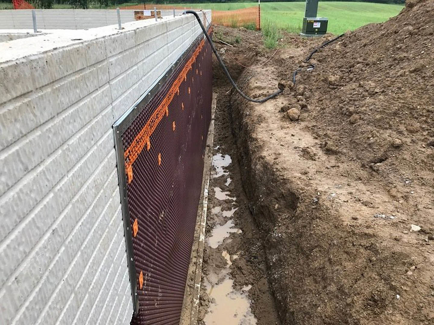 Excavated construction site with exposed soil, temporary fencing, and a visible trench containing a pipe; surrounding area includes grass and composite materials.