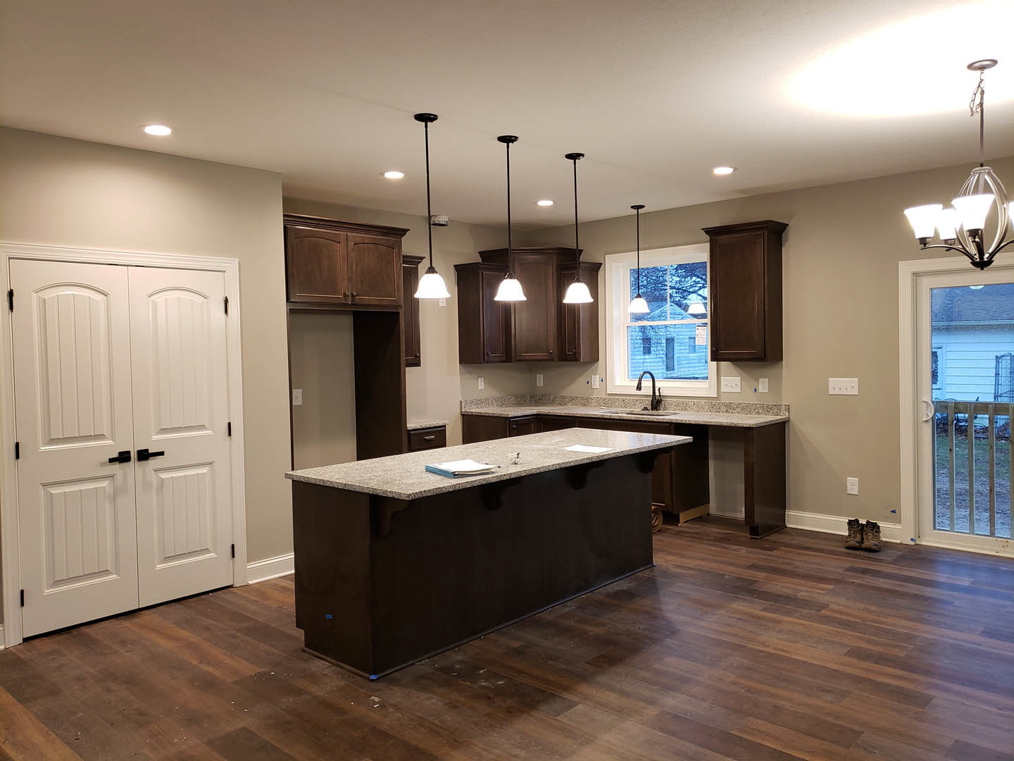 Spacious kitchen featuring a large central island with papers on the countertop, white double doors with black handles, brown cabinetry beneath a window, sliding glass door, and