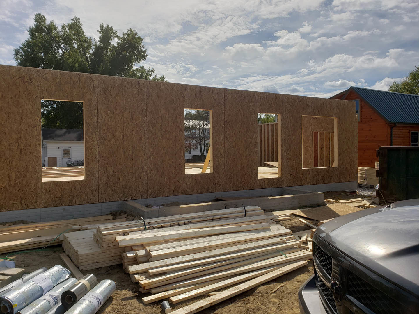 Partially built house with blue roof, exposed framing, stacked lumber, and a parked car on dirt driveway under a cloudy sky