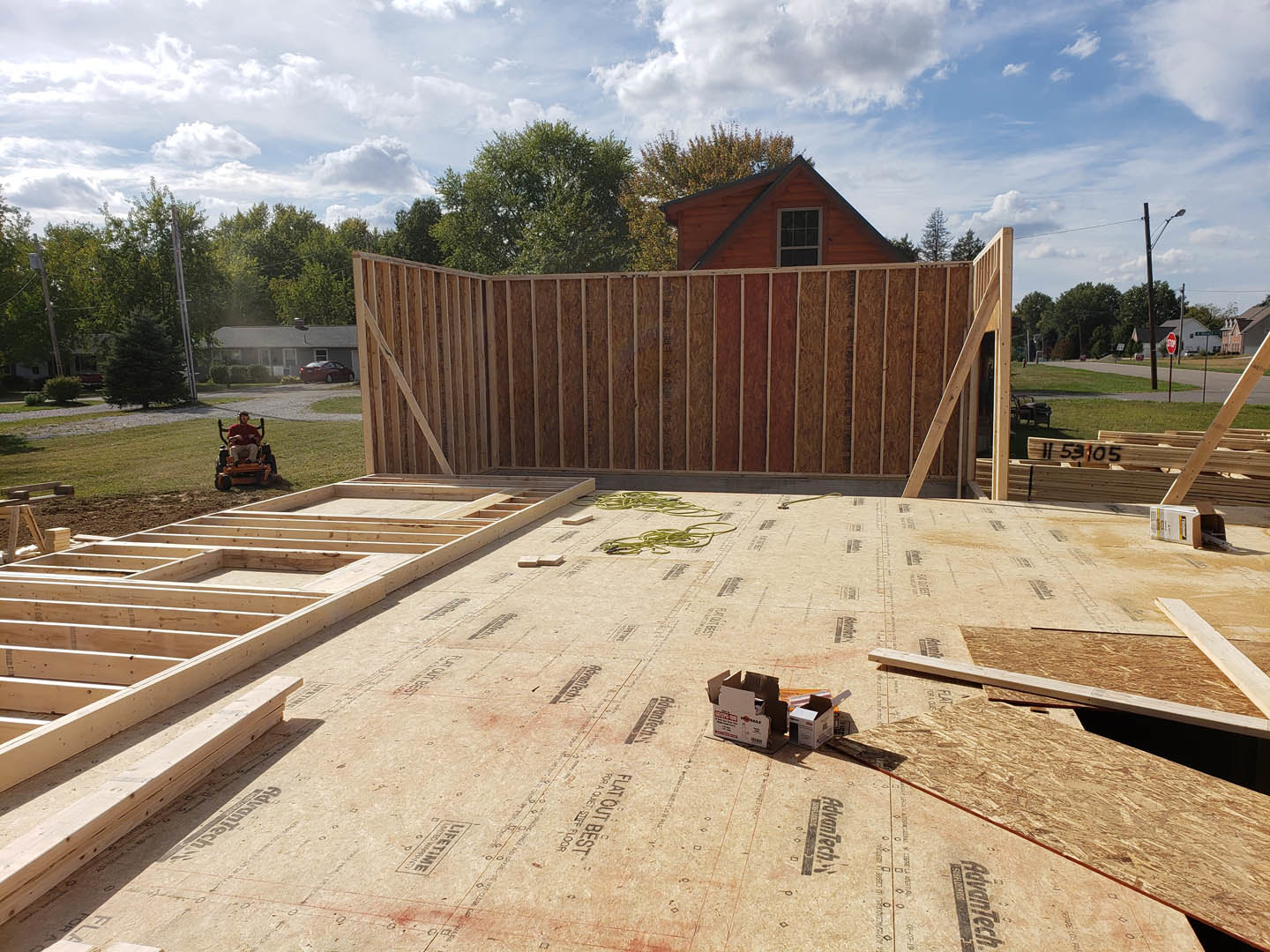 Wood-framed house under construction with exposed lumber, partially finished wood floor, grid window, wood paneled fence, boxes on a surface, and a man in a red shirt riding a