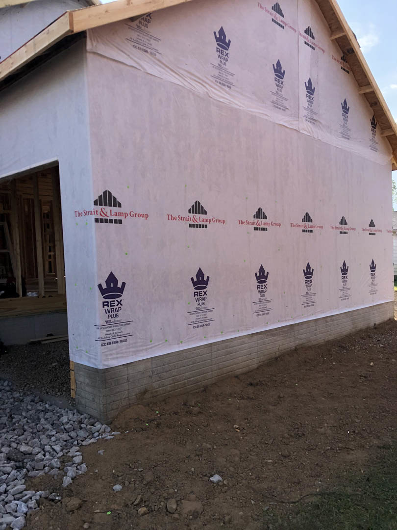 Partially built house with exposed wooden framing, white plastic sheeting draped over exterior wall, piles of rocks and dirt in foreground, unfinished roof structure visible under