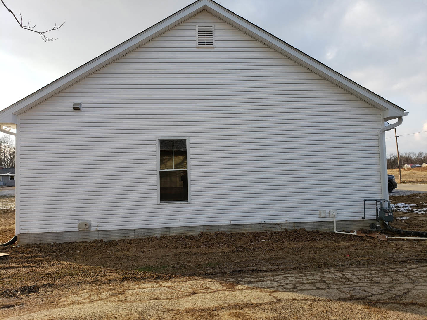 White siding house with single window, dirt road in foreground, tire tracks on soil, tree branch visible against cloudy sky