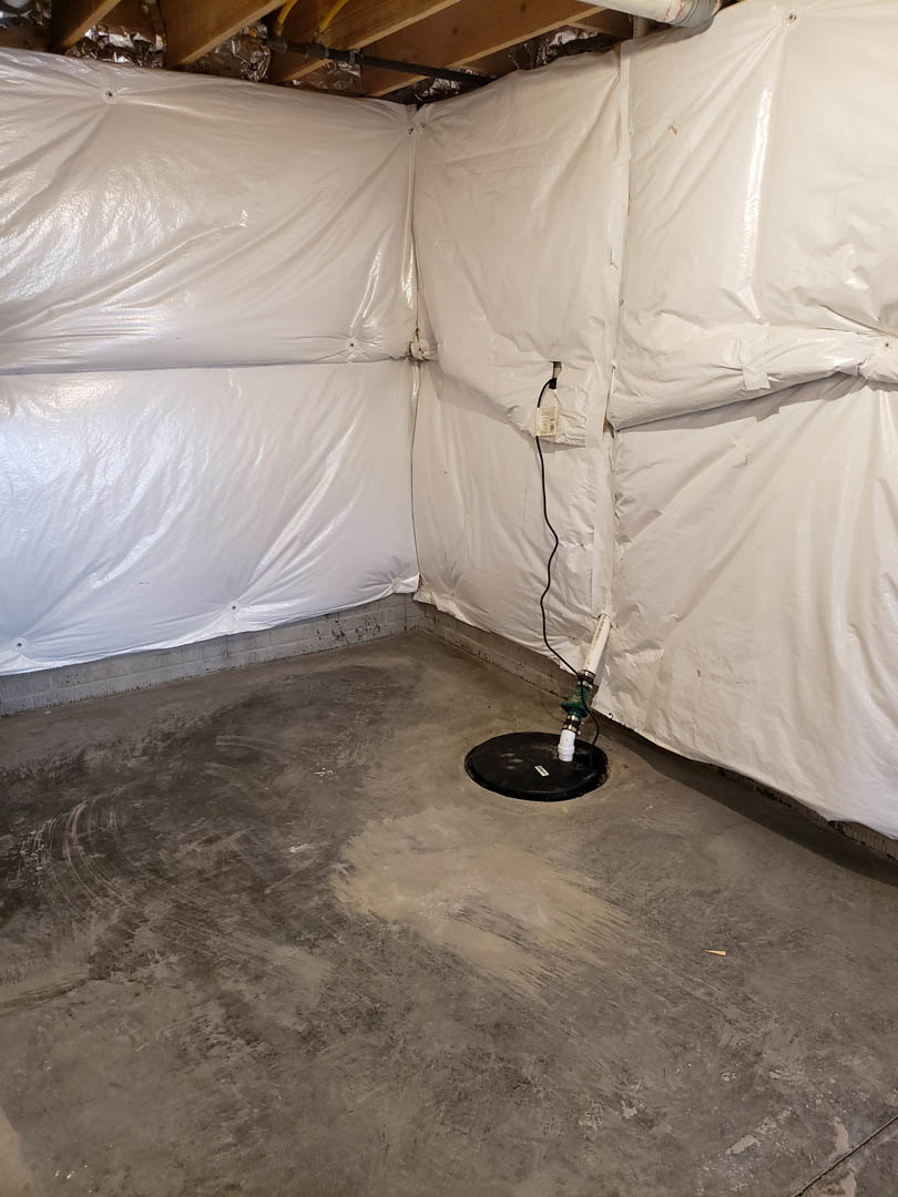 Basement with white plastic covering on walls and ceiling, concrete floor, black round object with attached white pipe, exposed wood ceiling with connected pipe, white tag hanging