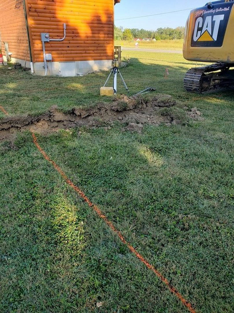 Excavated construction site with exposed dirt, grassy perimeter, parked vehicle, metal pipe against wooden wall, and clear sky overhead