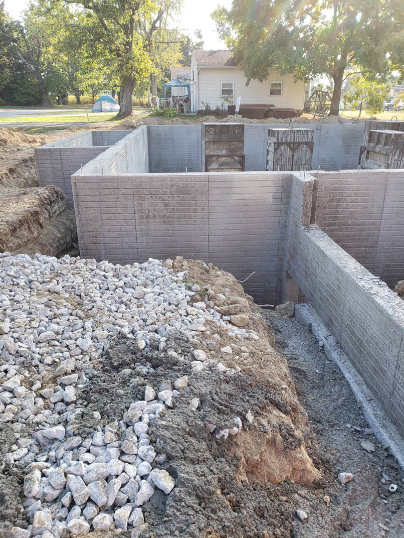 Concrete foundation surrounded by rocks and dirt, wooden box with metal frame in foreground, white house and trees visible in background