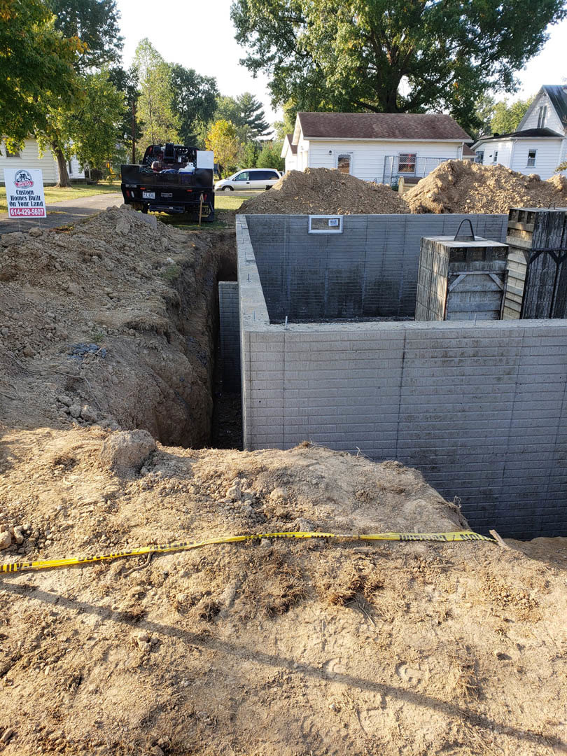 Excavated foundation surrounded by dirt, concrete retaining wall, yellow caution tape, black truck parked nearby, white house with fenced yard in background