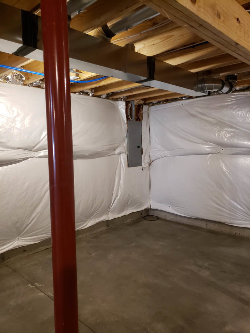 Basement with concrete floor, exposed metal beams and pipes overhead, white plastic sheeting covering walls and surfaces, red pipe and pillar visible.
