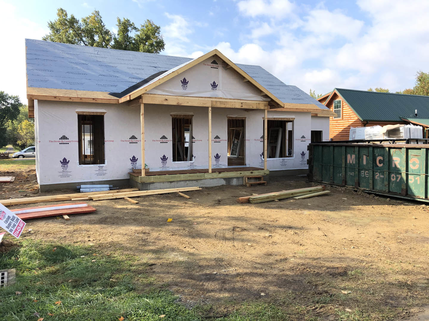 Framed house under construction with shingled roof, green dumpster with white writing, dirt yard scattered with wood pieces, temporary sign with black text, close-up of unfinished