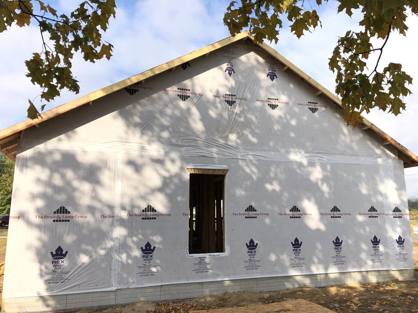Framed house under construction with exposed plywood walls, unfinished roof, and surrounding mature trees in the background