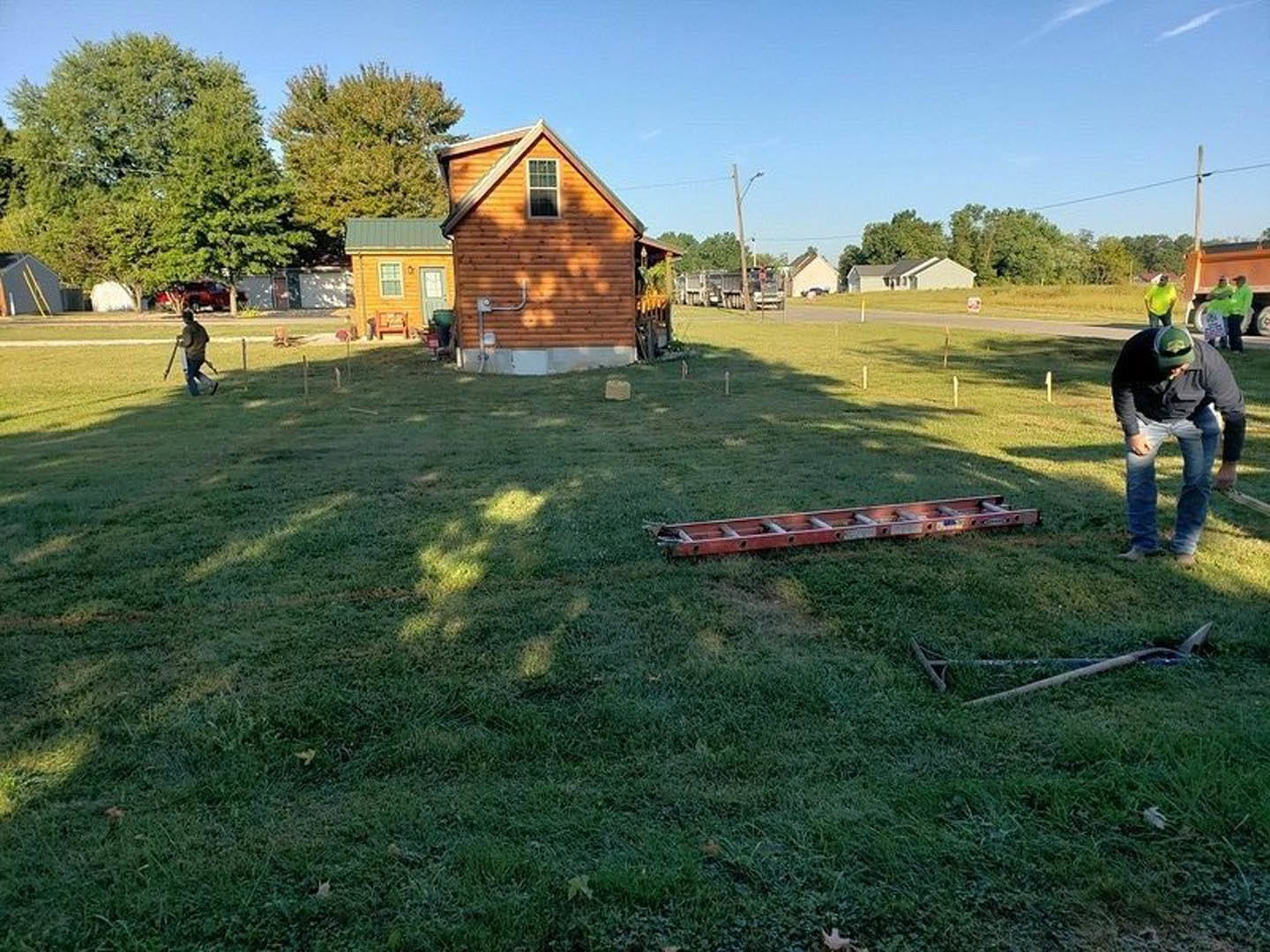 Modern two-story house with white siding and large windows, tree shadow cast on exterior wall, lush green lawn in foreground, man wearing green hat mowing grass, wooden ladder