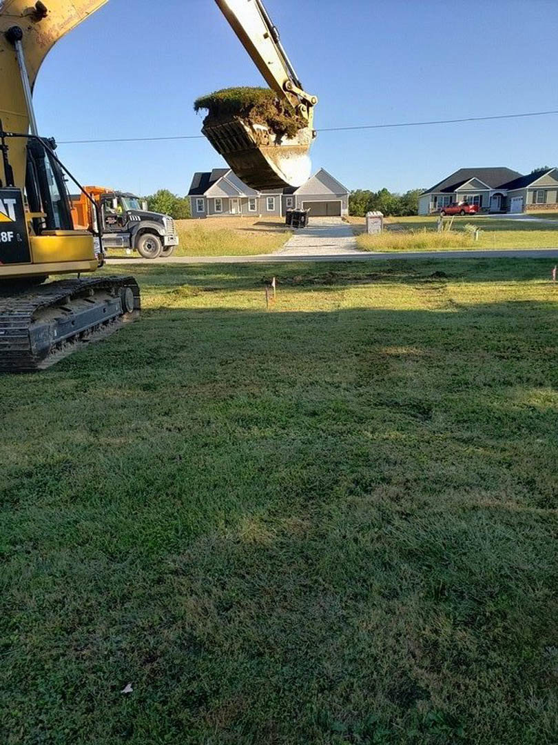 Yellow construction vehicle with caterpillar tracks parked on grassy field, bucket filled with green grass, trees and blue sky in background
