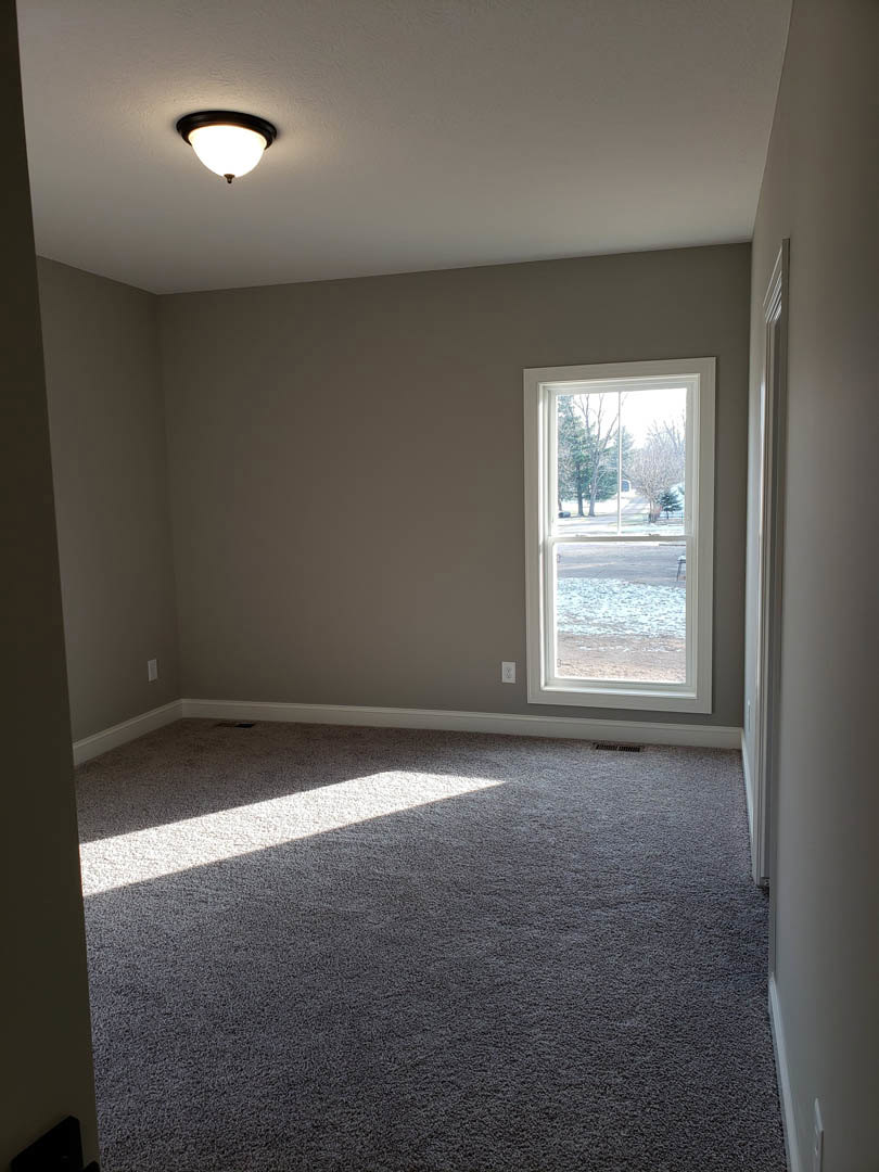 Carpeted room with white walls, ceiling light fixture, large window overlooking snowy yard and trees