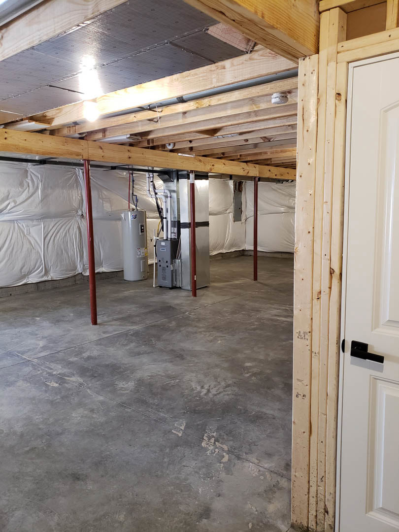 Room with exposed wood ceiling beam, white paneled door, concrete floor, red support poles, large white cylinder with black button, visible ceiling pipe and light fixture.
